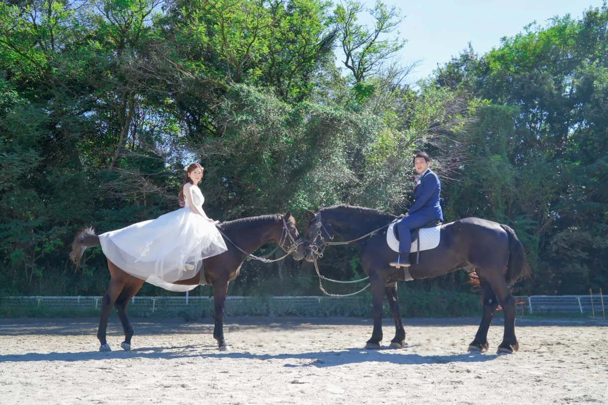 緑に囲まれた馬場で2頭の馬に乗って向かい合うウェディングドレスと青いタキシードのカップル