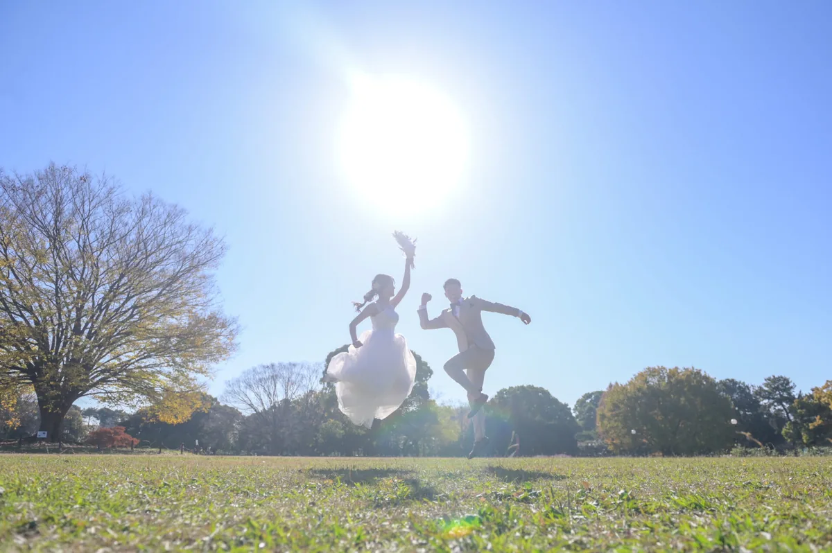 秋晴れの公園の芝生で太陽を背にジャンプするウェディングドレスとタキシードのカップル