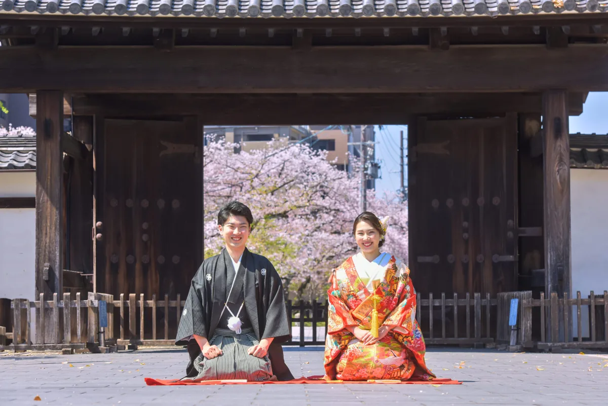 桜が咲く歴史的な門の前で色打掛と黒紋付袴で正座する和装の前撮り写真