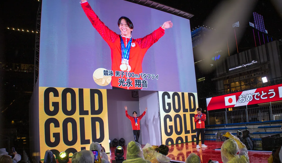 大型ビジョンに映し出された競泳男子100mバタフライ金メダルの光永翔音選手と、ランウェイで手を振る選手たち