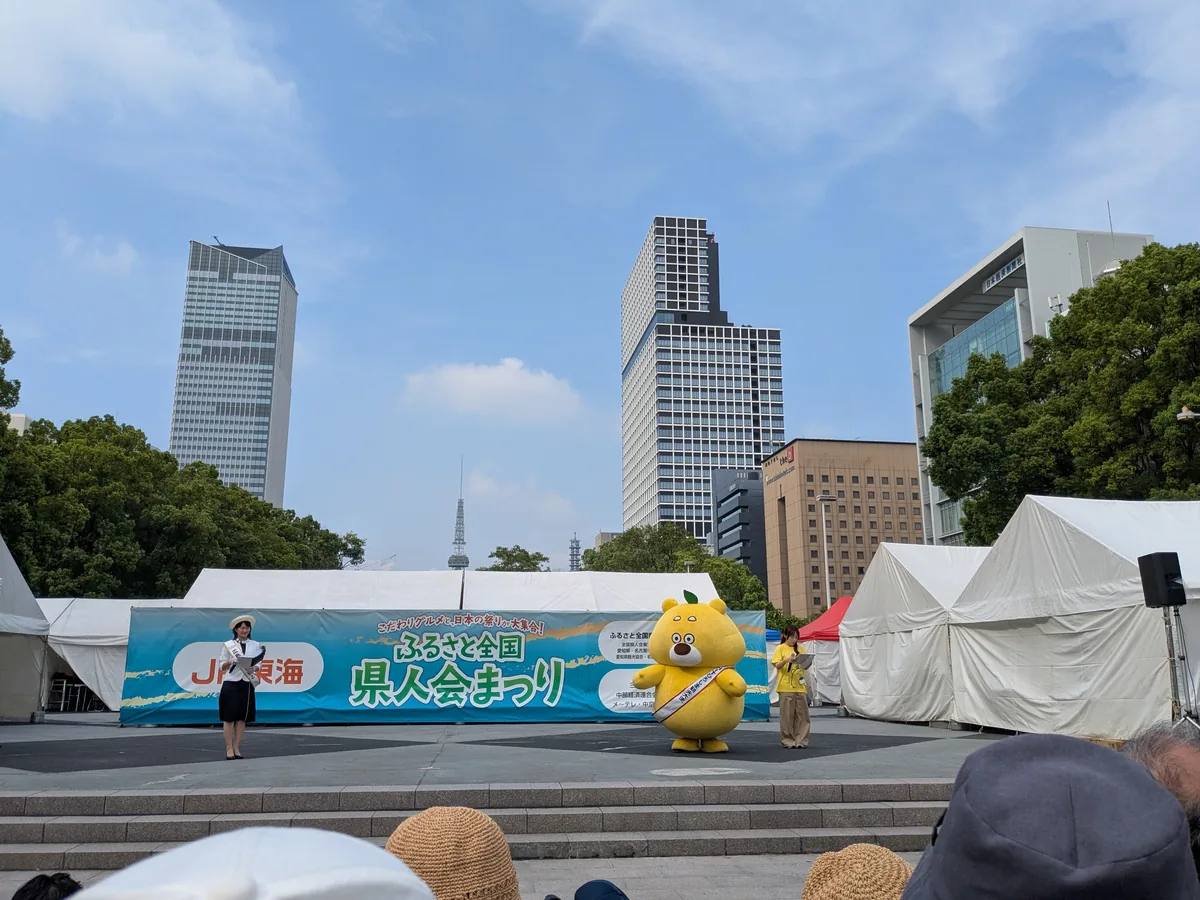 ふるさと全国県人会まつり2026のステージに登場した黄色いご当地キャラクターと司会者。背景に名古屋テレビ塔