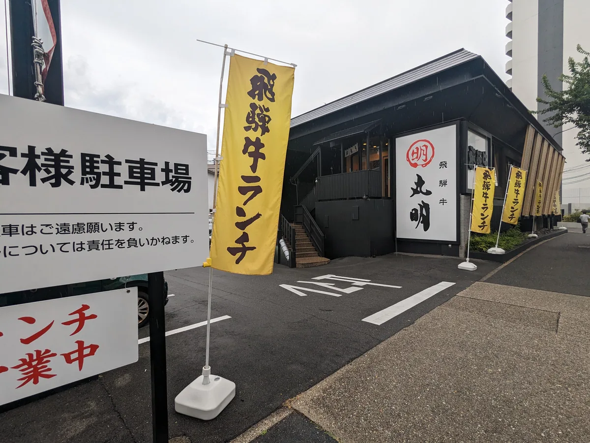 丸明 茶屋ヶ坂店の外観。黒い一軒家の店舗と飛騨牛ランチののぼり、お客様駐車場