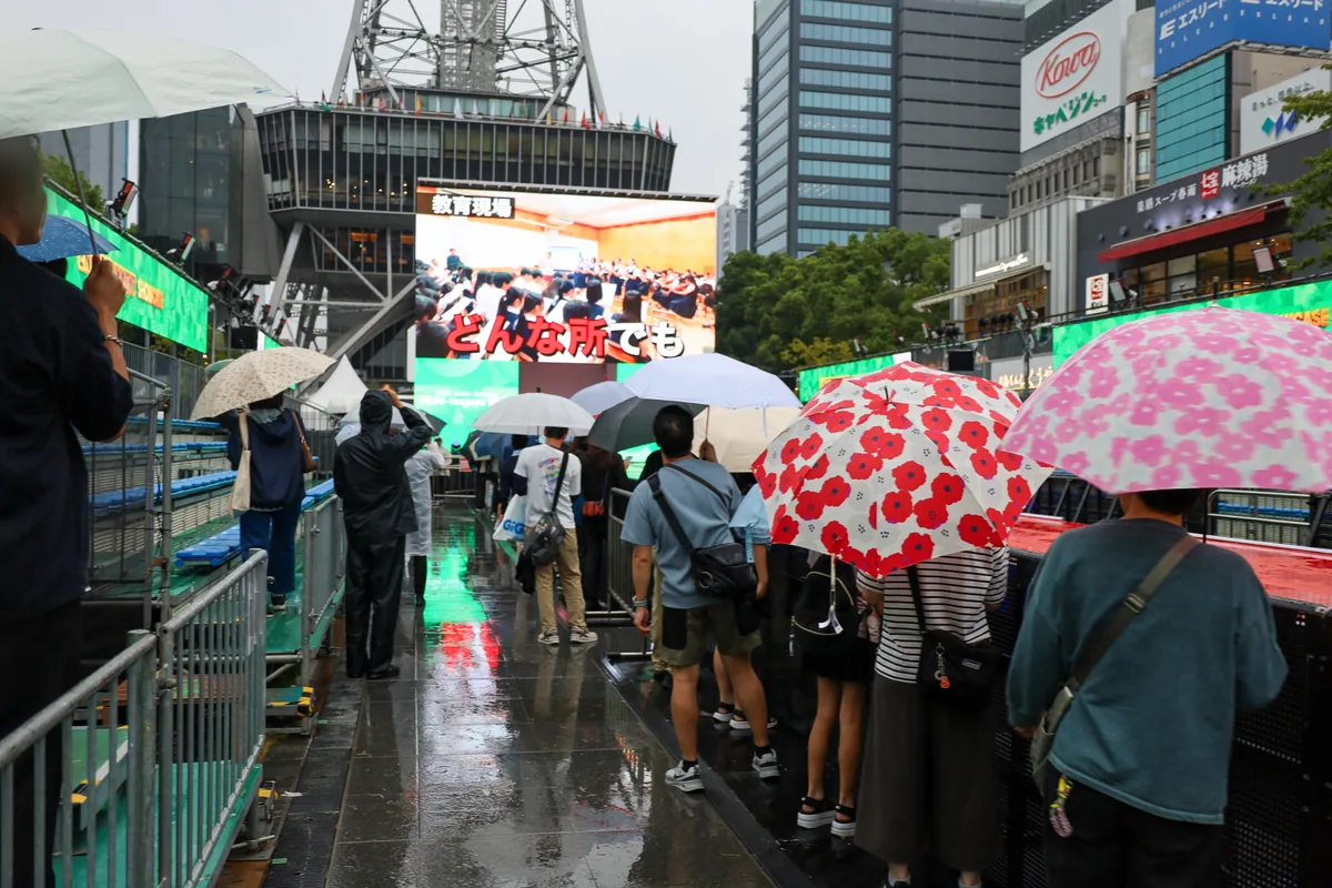 雨の中、傘をさして大型ビジョンのステージ映像を見る観客。9月26日のCHAMPIONS ROAD & STAGE