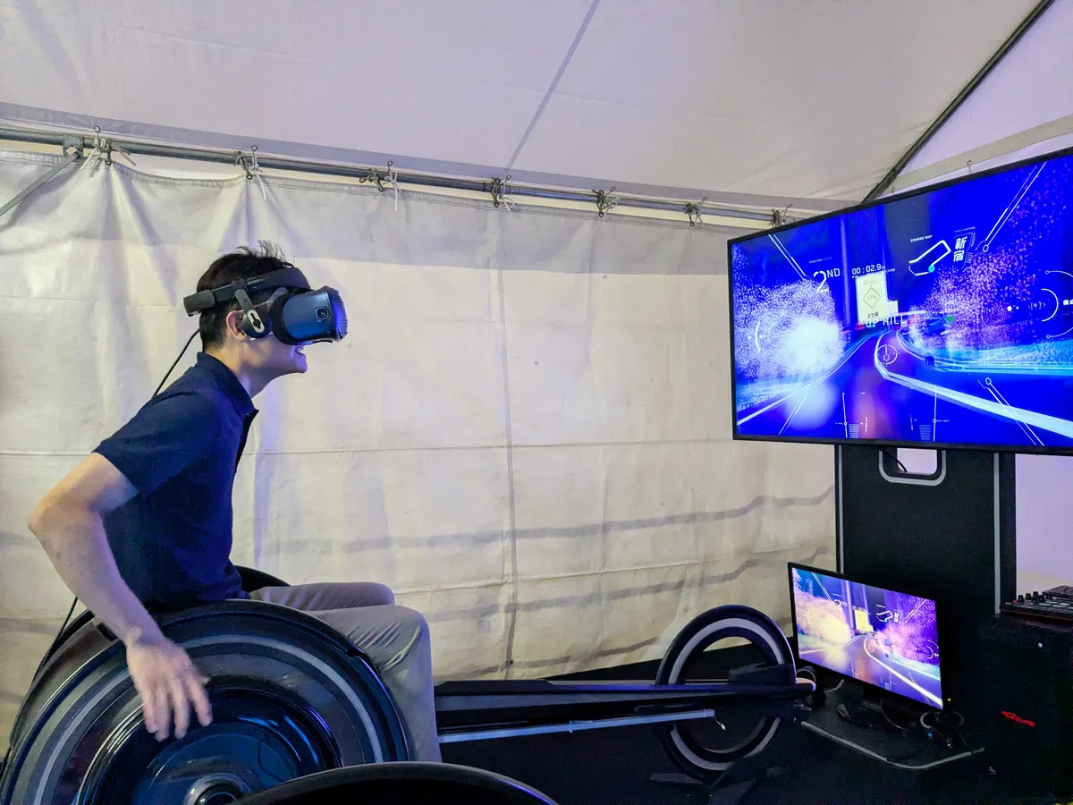 アジアパラ ライブサイトの車いすレースVR「CYBER WHEEL X」。VRゴーグルをつけた記者がレース用マシンの車輪を両手で回し、モニターに未来都市のコースが映っている