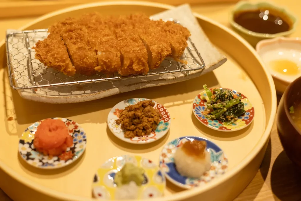 ロースかつ御膳〈豆皿仕立て〉の丸いお盆。網にのったロースかつの手前に、塩とわさび、豚のしぐれ煮、ポン酢ジュレをのせた大根おろしなど5つの豆皿が並ぶ
