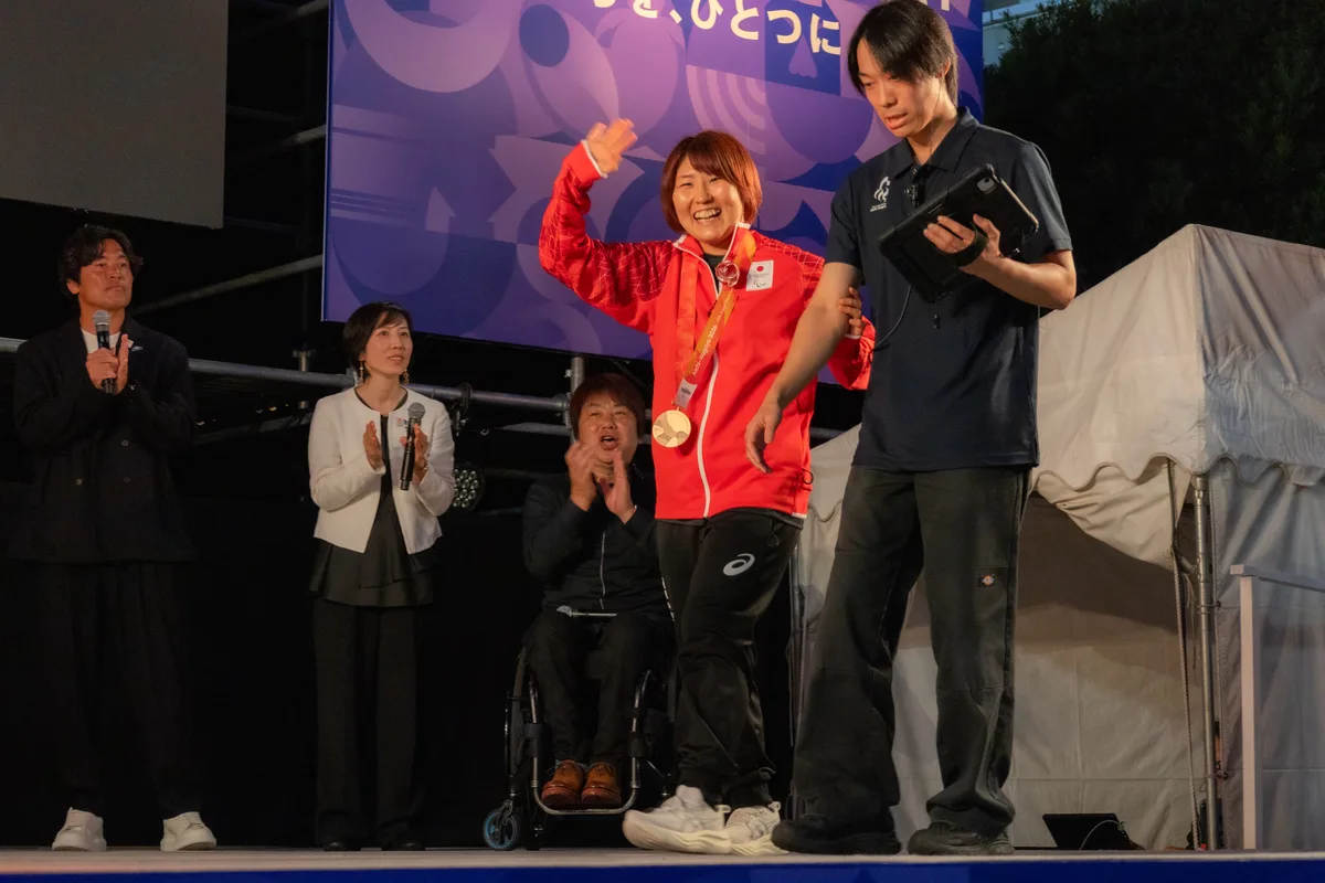銅メダルを掛けて笑顔で手を振るパラ柔道の藤原由衣選手。後ろで拍手する根木慎志さんと勅使川原郁恵さん