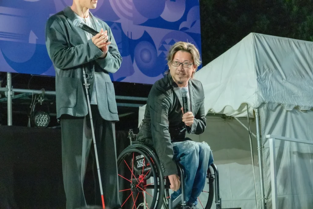 車いすでマイクを持って話す池崎大輔さんと、隣に立つ廣瀬誠さん