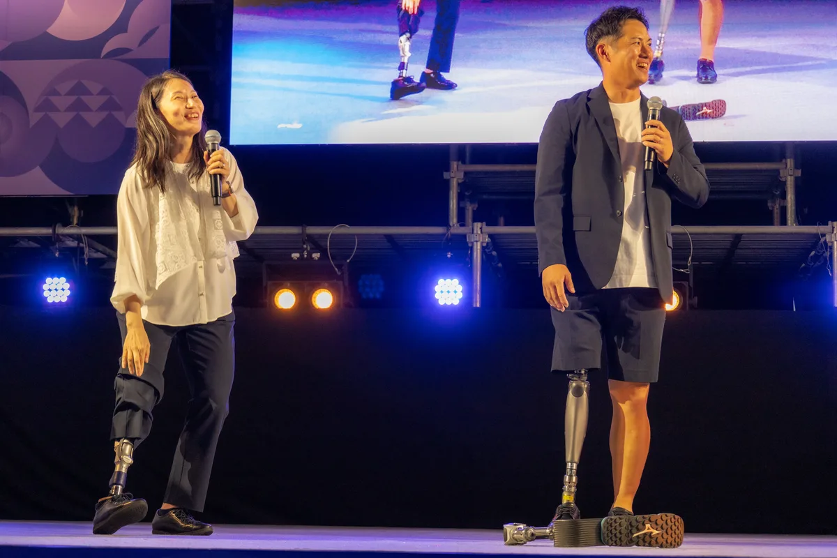 日常生活用の義足をステージで見せる小須田潤太さんと、隣で見る伊藤則子さん
