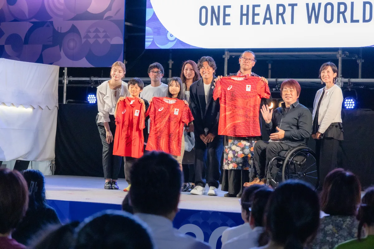 サイン入りの赤いチームジャパンのウェアを受け取った会場の3人と、レジェンドアスリートの4人がステージに並ぶ