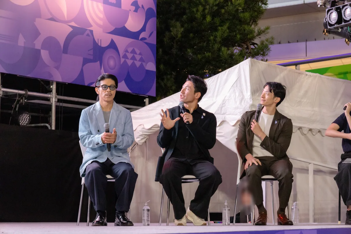 ステージの椅子に座り、開会式の中継を見ながら話す松本利夫さん、塚原直貴さん、入江陵介さん