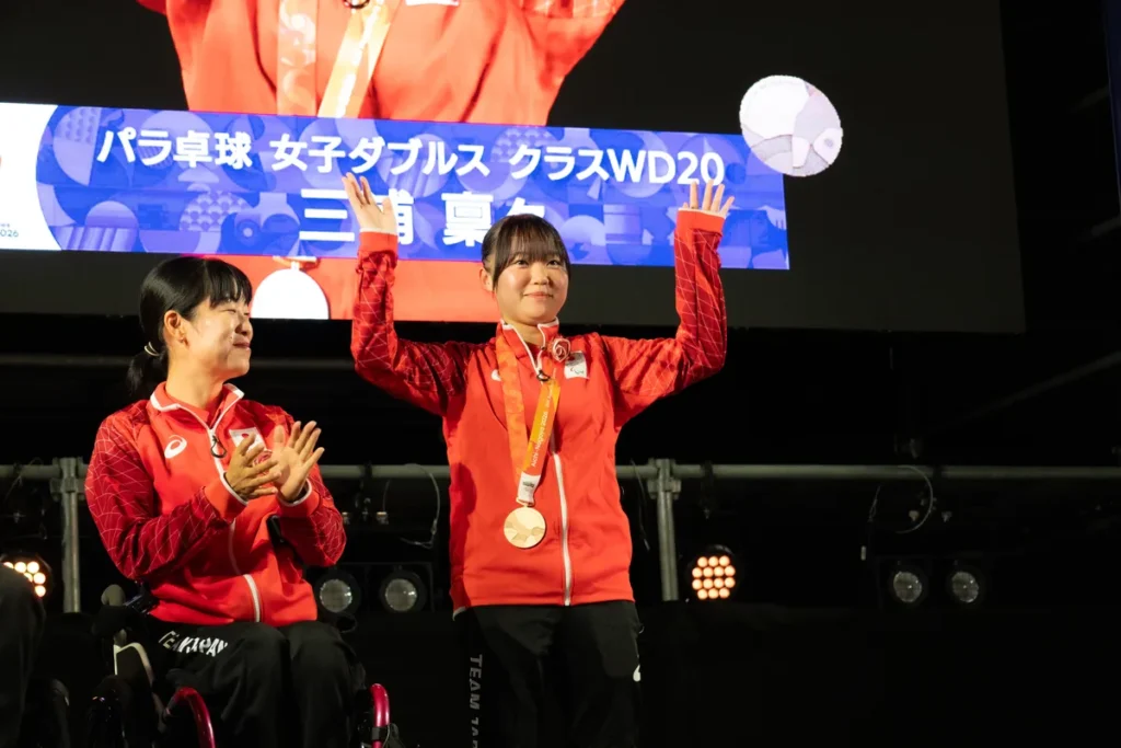 アスリートセレブレーションのステージで名前を呼ばれ、手を挙げるパラ卓球の三浦稟々選手。左はパラアーチェリーの篠原彩選手