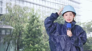 台風の雨の中、ヘルメットとレインウェア姿でマイクを持ち中継するリポーターのイメージ