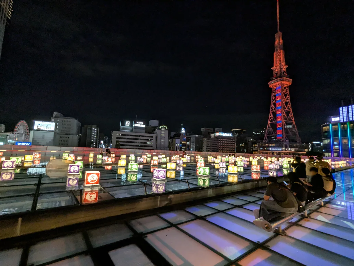 夜のオアシス21の屋上。水面に色とりどりの光るオブジェが並び、奥に赤くライトアップされた中部電力MIRAI TOWERと栄の街並み