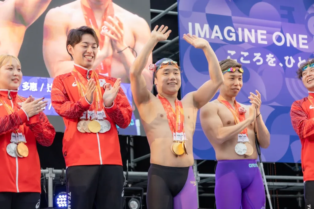 黒い競泳水着で登壇して両手を振る齋藤元希選手と、隣で拍手する富田宇宙選手