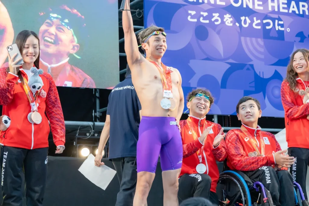 競泳の水着姿でステージに立ち、片手を上げる富田宇宙選手。隣でスマートフォンを構える辻内彩野選手