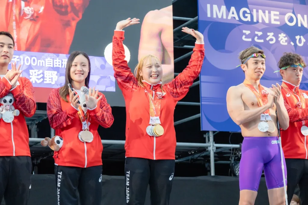 5つのメダルを下げて両手を振る辻内彩野選手と、隣で拍手する川辺多恵選手