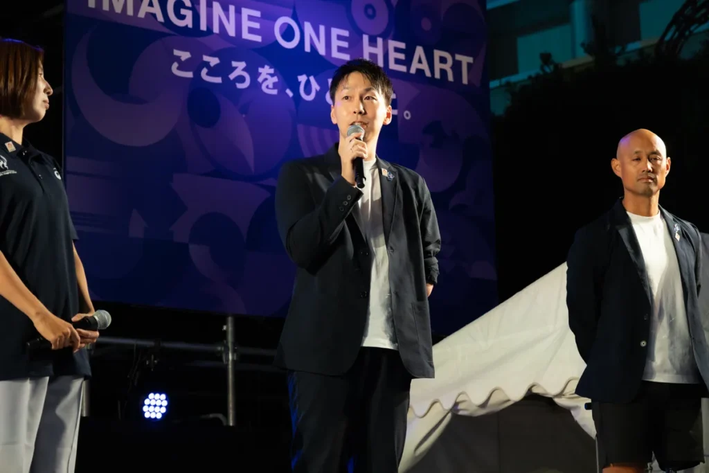 ONE HEART WORLDのステージでマイクを持って話す山田拓朗さんと、隣に立つ山本篤さん